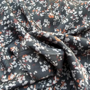 May include: Dark gray fabric with a floral pattern. The fabric features small white and orange flower-like shapes scattered across its surface. The fabric is draped, creating folds and shadows that add depth and texture to the design.