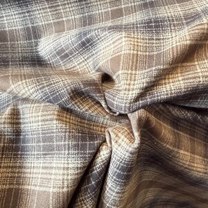 May include: Close-up of a fabric with a plaid pattern. The fabric features a blend of brown, gray, and off-white colors. The plaid design is composed of intersecting lines, creating a classic and textured appearance. The fabric is folded, showing the texture.