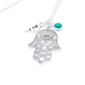 Può includere: Collana d'argento con un ciondolo a forma di mano di hamsa, un piccolo ciondolo con una lettera e una pietra preziosa verde. Il ciondolo a forma di mano di hamsa ha un occhio al centro e dettagli intricati.