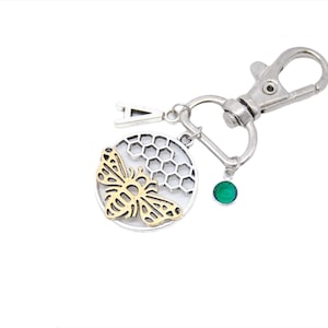 Puede incluir: Llavero plateado con un dije de abeja dorada, un diseño de panal, un dije de gema verde y un dije de letra A.