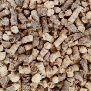 May include: Close-up of a pile of brown and tan wood pellets. The pellets are cylindrical and have a rough, textured surface. The image is well-lit, showing the details of the wood grain and the varying shades of brown.