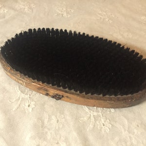 Könnte beinhalten: Ovale, antike Kleiderbürste mit Holzrahmen und schwarzen Borsten. Die Bürste ist abgenutzt, mit sichtbaren Gebrauchsspuren. Der Holzrahmen hat eine hellbraune Farbe.