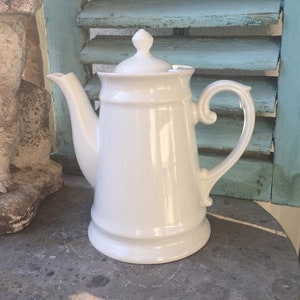 Könnte beinhalten: Eine weiße Keramik-Kaffeekanne mit Deckel und gebogenem Griff. Der Topf steht auf einer grauen Steinoberfläche, im Hintergrund eine hellblaue Jalousie. Die Kanne hat ein klassisches Design.