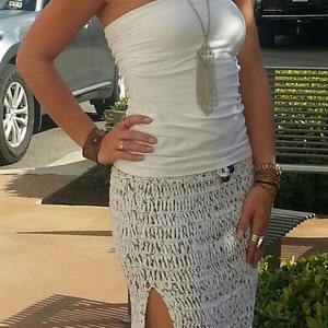 May include: A white crochet maxi skirt with a side slit. The skirt is paired with a white strapless top and a silver chain necklace with a tassel pendant.