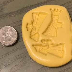 May include: A light yellow, irregular-shaped mold with three detailed figures of kings, each holding a gift. A US quarter is next to the mold for size comparison. The mold is likely for making decorative items.