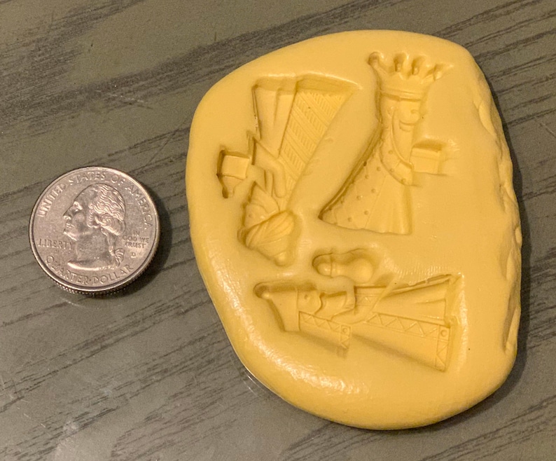 May include: A light yellow, irregular-shaped mold with three detailed figures of kings, each holding a gift. A US quarter is next to the mold for size comparison. The mold is likely for making decorative items.