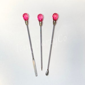 May include: Three stainless steel tools with pink faceted crystal tops. One tool has a flat, rectangular end, another has a pointed end, and the third has a rounded, spoon-like end. The tools are arranged on a white surface.