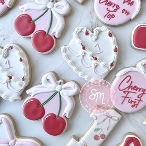 Puede incluir: Surtido de galletas de azúcar decoradas con temática de cerezas. Las galletas incluyen cerezas, lazos y formas de corazón, con las palabras "Cherry on Top" y "Cherry First" en glaseado rojo. Las galletas son rosas, blancas y rojas.