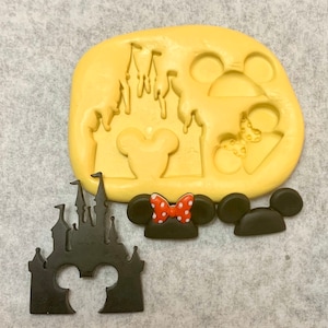 Peut inclure: Un moule en silicone jaune avec des motifs de Mickey Mouse et de château, ainsi qu'une découpe de château noire et deux formes d'oreilles de Mickey Mouse noires, dont une avec un nœud rouge à pois blancs.