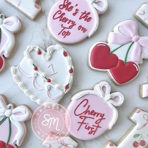May include: Assortment of decorated sugar cookies with a cherry theme. Cookies include pink and white icing with cherry and bow designs. Some cookies have text that reads "She's the Cherry on Top" and "Cherry First".