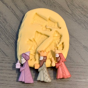 May include: A set of three miniature Wise Men figurines in purple, green, and pink robes, each holding a gift or lantern. A yellow mold with Wise Men figures is in the background. The figurines are likely for a nativity scene.