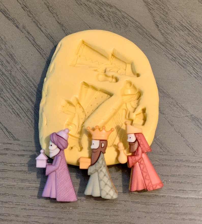 May include: A set of three miniature Wise Men figurines in purple, green, and pink robes, each holding a gift or lantern. A yellow mold with Wise Men figures is in the background. The figurines are likely for a nativity scene.