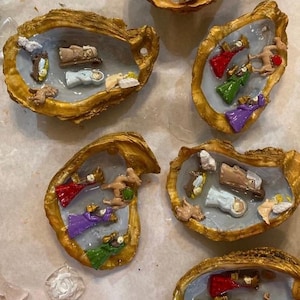May include: Several oyster shells, each containing a miniature nativity scene. The shells have a gold-colored exterior and a light blue interior. The nativity figures include baby Jesus, angels, and the three wise men.