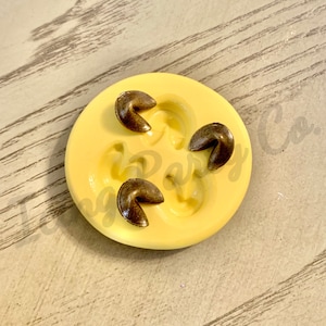 Puede incluir: Un molde de silicona amarillo con tres galletas de la fortuna de color bronce. El molde tiene hendiduras para las galletas. La imagen está sobre una superficie clara con vetas de madera.