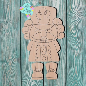May include: A wooden cutout of a nutcracker soldier with a smiling face, curly hair, and a tall hat. The nutcracker has a coat with three buttons and boots. The background is a teal-colored wooden surface. The image includes a logo that says "Kitsch & Confetti".