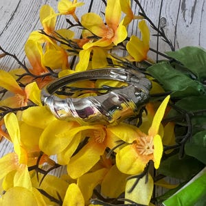 May include: A silver twisted bangle bracelet with a hinged clasp. The bracelet is surrounded by yellow silk flowers.