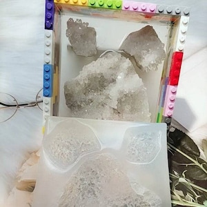 May include: A clear plastic box with a colorful Lego border contains a large cluster of white quartz crystals. A ruler is placed below the box, showing measurements in inches.