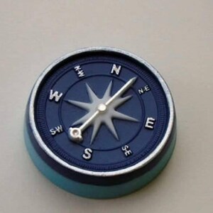 Op de afbeelding: Een blauwe kompas met een zilveren naald die naar het noorden wijst. Het kompas heeft een witte ster in het midden en is omgeven door een zilveren ring met de windrichtingen in zwart.