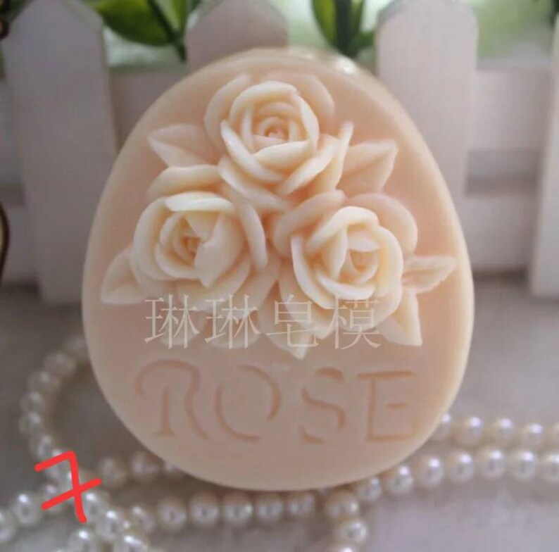 May include: A pale pink oval-shaped soap with three raised roses and the word "ROSE" embossed on the front. The soap is sitting on a white surface with a string of white pearls.