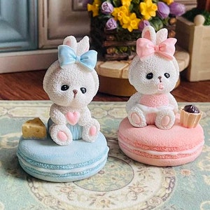 Puede incluir: Dos figuritas de conejito blanco con lazos rosas y azules sentados encima de macarons rosa pastel y azul. El conejito de la izquierda sostiene un trozo de queso y el conejito de la derecha sostiene un pequeño cupcake de chocolate.