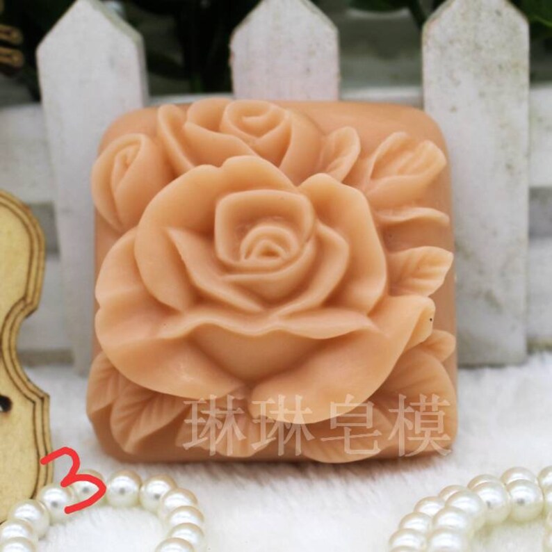 May include: A square-shaped silicone mold with a detailed rose design. The mold is made of a light pink silicone and is perfect for making soap, candles, or other crafts.