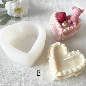 May include: A white silicone heart-shaped mold for making candles. The mold has a smaller heart-shaped cavity inside. A finished heart-shaped candle with white frosting and a pink heart-shaped cake with a strawberry and a pink teddy bear on top are shown.