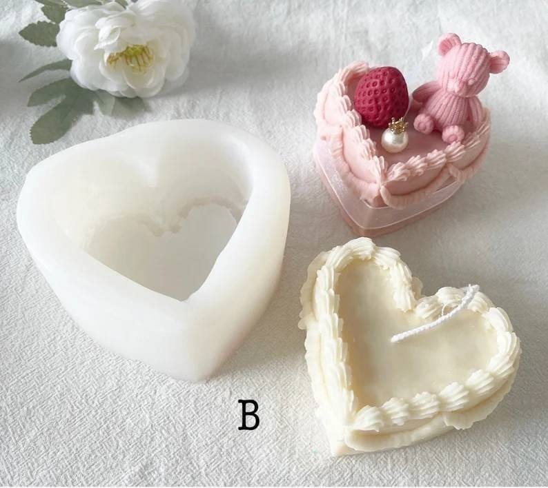May include: A white silicone heart-shaped mold for making candles. The mold has a smaller heart-shaped cavity inside. A finished heart-shaped candle with white frosting and a pink heart-shaped cake with a strawberry and a pink teddy bear on top are shown.