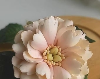 May include: A delicate, pale pink flower with gold accents, crafted from polymer clay. The flower has a layered, ruffled appearance and is set against a wooden background.