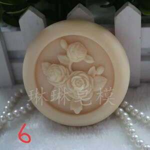 May include: A round, off-white soap with a raised design of three white roses. The soap is surrounded by a white border and the text "森林 皂 芙" is printed in black on the soap.