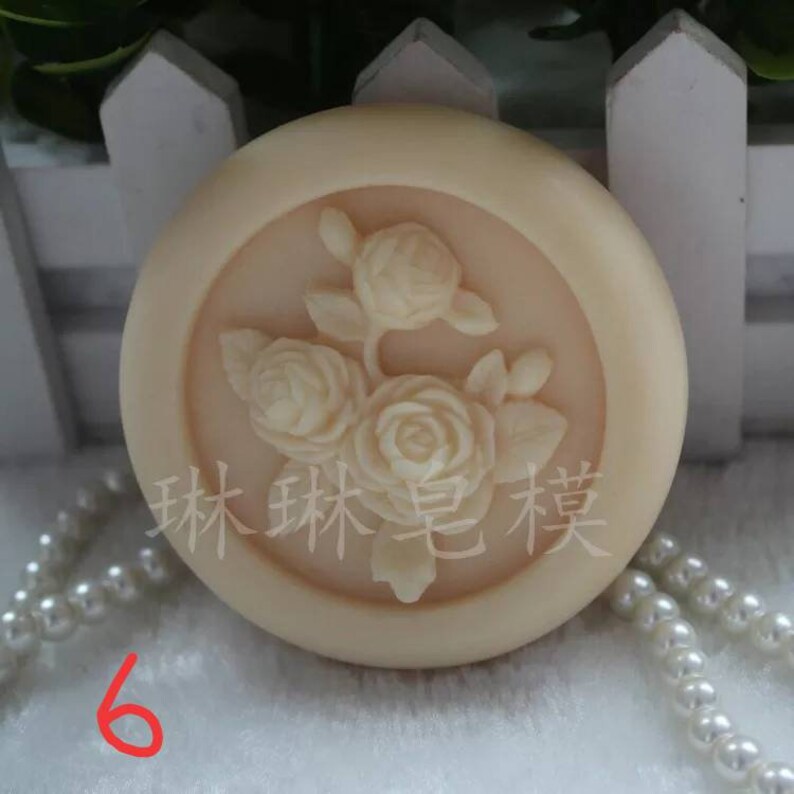 May include: A round, off-white soap with a raised design of three white roses. The soap is surrounded by a white border and the text "森林 皂 芙" is printed in black on the soap.