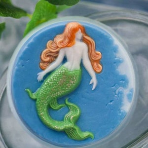 Op de afbeelding: Een ronde, blauwe en witte zeep met een zeemeerminontwerp. De zeemeermin heeft lang, rood haar, een groene staart en een witte torso.