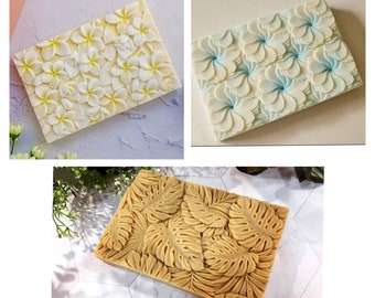 May include: Three rectangular soap bars with different designs. The first bar has a white background with a raised design of white frangipani flowers. The second bar has a light blue background with a raised design of light blue frangipani flowers. The third bar has a light brown background with a raised design of brown monstera leaves.
