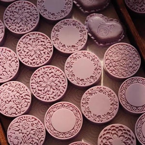 Puede incluir: Una colección de jabones redondos de color púrpura con intrincados diseños florales y geométricos. Los jabones están dispuestos en una caja de madera.