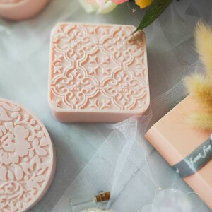 K&ouml;nnte beinhalten: Zwei rosa Seifenst&uuml;cke mit komplizierten Blumenmustern. Die Seifenst&uuml;cke liegen auf einem wei&szlig;en T&uuml;llstoff-Hintergrund.