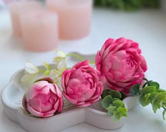 May include: Three pink peony flower buds on a gray tray with green leaves. The flowers are arranged in a cluster, with the largest bud in the center and the two smaller buds on either side. The flowers are made of a soft, pliable material, possibly fondant or gum paste.