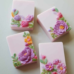 May include: Four rectangular bars of soap with floral designs. Each bar is a light pink color and features a different floral design in shades of purple, pink, yellow, and green.