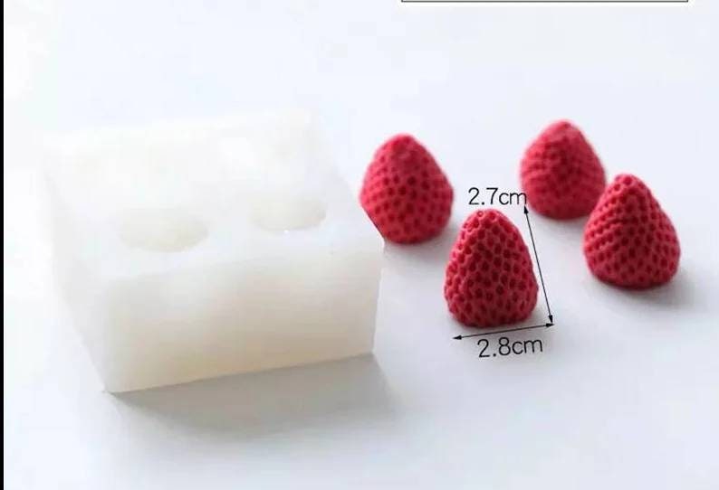 May include: A white silicone mold with four cavities shaped like strawberries. Four red strawberry-shaped candies are next to the mold. The strawberries are 2.7 cm wide and 2.8 cm tall.