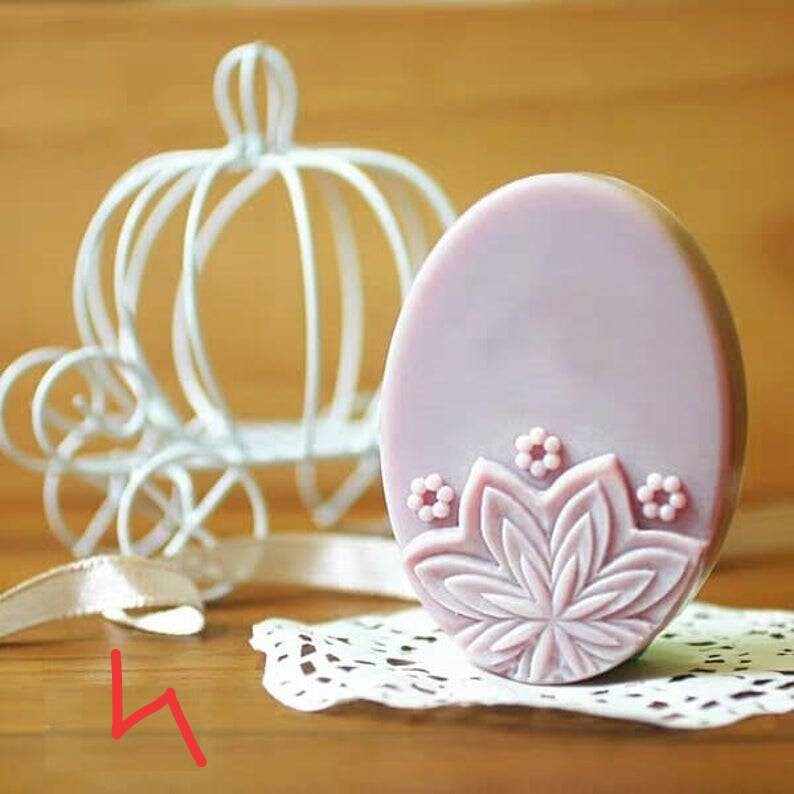K&ouml;nnte beinhalten: Eine runde, helllavendelfarbene Seife mit einem detaillierten Blumenmuster. Die Seife hat ein erhabenes, gepr&auml;gtes Design einer Blume mit einer Mitte und Bl&uuml;tenbl&auml;ttern.
