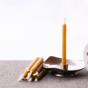 Op de afbeelding: Een enkele bijenwaskaars in een zilveren houder met een groep kleinere bijenwaskaarsen op een grijs oppervlak. Een witte keramische cilinder met gaten bevindt zich op de achtergrond.
