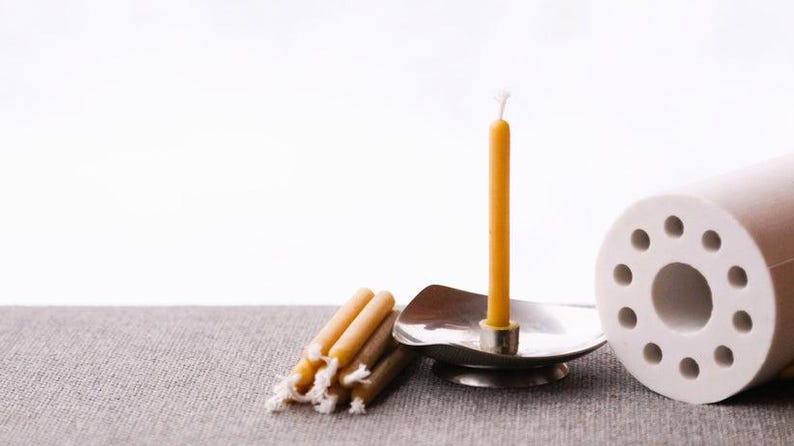 Op de afbeelding: Een enkele bijenwaskaars in een zilveren houder met een groep kleinere bijenwaskaarsen op een grijs oppervlak. Een witte keramische cilinder met gaten bevindt zich op de achtergrond.