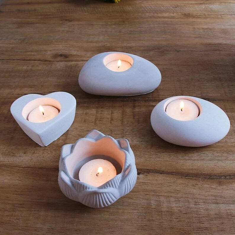 May include: Four white concrete candle holders in different shapes: a heart, a pebble, a flower, and another pebble. Each holder has a small tea light candle inside, with the flames lit.