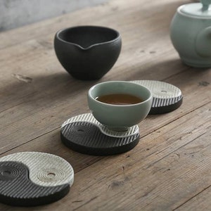 May include: Three black and white yin yang ceramic coasters with a small light green teacup and saucer on one of the coasters. The teacup is filled with brown tea. A light green teapot is in the background.