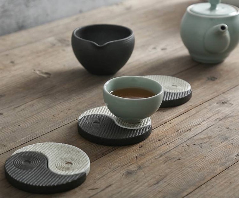 May include: Three black and white yin yang ceramic coasters with a small light green teacup and saucer on one of the coasters. The teacup is filled with brown tea. A light green teapot is in the background.