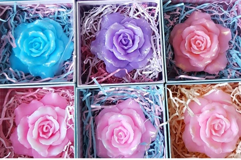 May include: Six small boxes each containing a single rose-shaped soap. The soaps are in shades of pink, light blue, and purple.