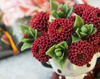 May include: A bouquet of red felt flowers with green felt leaves in a white ceramic vase with a cartoon face.