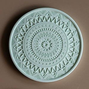May include: A round, light green coaster with a raised, intricate geometric design. The design features concentric circles of triangles, diamonds, and other shapes.