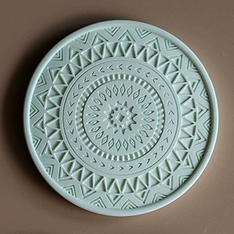 May include: A round, light green coaster with a raised, intricate geometric design. The design features concentric circles of triangles, diamonds, and other shapes.