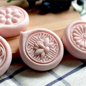 May include: Four pink soap bars with different floral designs. The soaps are on a wooden surface with a checkered tablecloth.