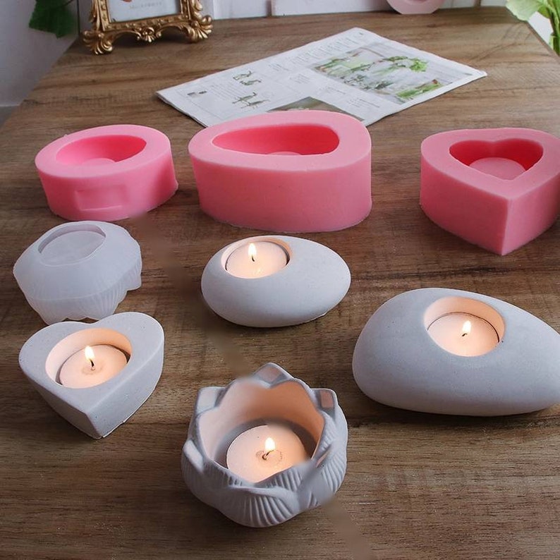 May include: Assortment of pink and gray silicone molds for making candles. The molds are shaped like a heart, oval, flower, and a pebble. The molds are shown with finished concrete candle holders.