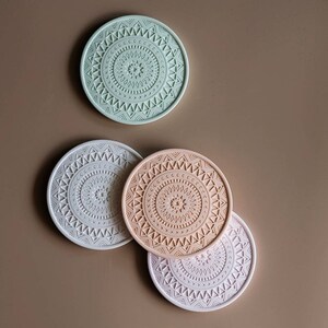 May include: Four round silicone coasters featuring a mandala design. The coasters are in shades of green, peach, and white.